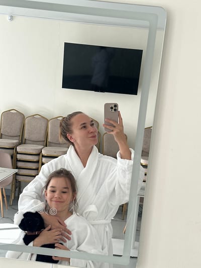 In a well-lit room, a woman and a young girl are reflected in a large mirror, posing for a selfie. Both are wearing white terrycloth bathrobes. The woman, with her hair pulled back in a bun, holds a smartphone in her right hand, capturing the reflection. Her left arm is around the girl's shoulders. The girl, to the left of the woman, is holding a small black and white stuffed animal. She is smiling, and her head is slightly tilted. The background visible in the mirror shows stacks of beige chairs with gold frames, a white table with a chair, and a black television mounted on a light-colored wall. A faint crack is visible on the wall above the television. The overall scene suggests a relaxed atmosphere, possibly in a hotel or spa. The image does not contain any recognizable landmarks or elements that would definitively place it in Terskol, Russia.
