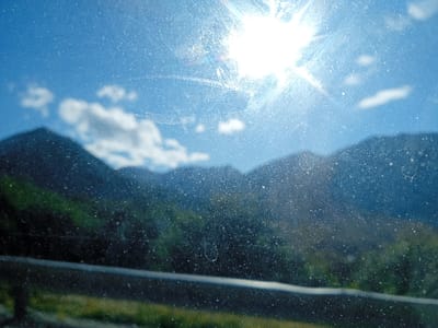 A landscape view is seen through a dirty window. A bright sun shines against a blue sky with a few clouds. Mountains rise in the distance, their lower slopes covered in green vegetation. A horizontal railing spans across the bottom of the image, obscuring some grass. The window is covered in dust, water spots, and scratches, which obscure the view.