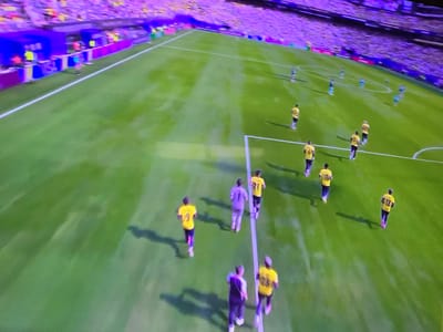 An elevated, wide-angle shot captures a football match in progress on a bright green, well-maintained pitch. Several players, most wearing yellow jerseys with black numbers and purple shorts, are dispersed across the field. Some are in motion, jogging or running, while others appear to be stationary. The spectators in the background are blurred, but their presence and the stadium seating are evident, bathed in a vibrant purple light. A white line marks the boundary of the playing field. The scene is likely within the city of Alpharetta, USA, given the context.