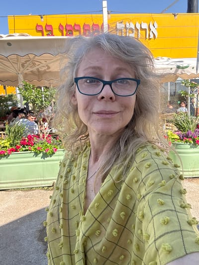 This is a medium shot of a woman with gray hair wearing glasses and a lime green patterned shirt, taken outdoors. She is looking towards the camera with a neutral expression. Behind her, a yellow sign with white lettering is visible. To her right, there is a green wall and an outdoor cafe setting with potted plants and flowers. The sky is clear and blue, suggesting a sunny day. The image is oriented vertically, with the woman's face on the left side. There are other people and umbrellas visible in the background, indicating a public outdoor space. The overall impression is casual and relaxed. The location is Bnei Shimon Regional Council, Israel.