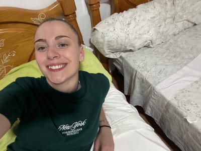 A young woman with blonde hair pulled back is lying on a bed, facing the camera and smiling. She is wearing a dark green t-shirt with white lettering that says "New York Five Boroughs MCMXCII". She has a bright yellow pillow behind her. To her right, the bed is made with white sheets and a duvet cover with a delicate floral pattern. The bed frame is made of light-colored wood with decorative carvings. The background shows a portion of a wooden headboard. The scene appears to be indoors, likely in a bedroom.