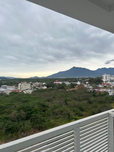 This is a wide-angle, vertical, eye-level shot of a cityscape and mountainous landscape, taken from a balcony in Palhoça, Brazil. The foreground features a white slatted railing of a balcony, partially obscuring the view. Beyond the railing, a dense green forest covers the middle ground, with scattered residential buildings and some taller structures with white facades. In the background, a range of dark, hazy mountains dominates the horizon under a cloudy, overcast sky. The light suggests it is late afternoon or early morning.