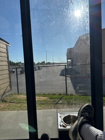 The image depicts a view from inside a structure, looking out through a glass pane that exhibits dirt and lens flare from the bright sun. The foreground shows a person's feet, one resting on a metal ashtray containing cigarette butts, and the other partially visible. The ashtray is positioned on a concrete surface. Beyond the glass, a chain-link fence encloses a paved area, likely a parking lot, with several cars parked in rows. A building, possibly a commercial establishment, is visible in the background, as are streetlights and trees. The sky is clear and blue. The location is the city of Anchorage, United States of America.
