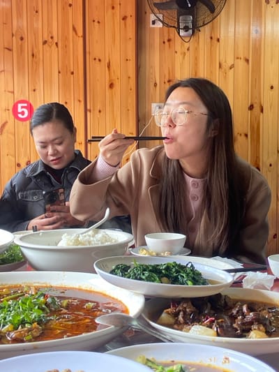 Two Asian women sit at a table laden with food. One woman, wearing glasses and a tan coat, eats with chopsticks. The other, in a black jacket, looks towards the food. Dishes of rice, stir-fried greens, meat, and fish are prominently displayed. A ceiling fan is visible in the background against a wooden wall. A red number '5' is at the bottom of the image.