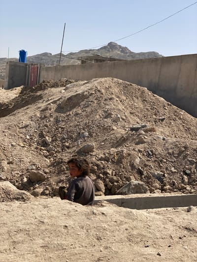 A child peers out from a small space between a large, coarse rock and a concrete wall. The child wears dark clothing and has dirty cheeks. Beyond the rock and wall, a structure is built into the side of a rocky mountain. A blue tank is visible in the distance. A thin wire stretches across the clear sky.