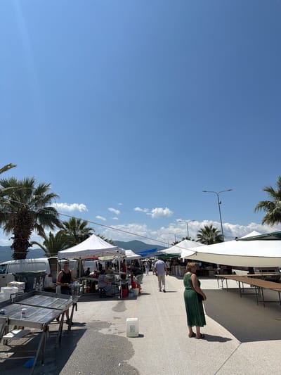 The image depicts an outdoor market on a sunny day, likely in Asprovalta, Greece, given the visual cues of palm trees, a clear blue sky, and mountains in the distance. In the foreground, a man stands near a metallic table, holding a bottle of water. To his right, a woman in a green dress is visible. The market features numerous stalls with white canopies, some of which are empty and set up with tables, while others are occupied with vendors and shoppers. Several white vans are parked behind the stalls. Palm trees are scattered throughout the scene, and streetlights are also present. The ground is paved with asphalt and concrete. The overall impression is that of a bustling marketplace during the daytime.