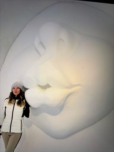 The image displays the interior of the Icehotel in Jukkasjärvi, near Kiruna, Sweden. A female subject is positioned in the lower-left quadrant, posing next to a large, white ice sculpture depicting abstract, puckered lips. The female wears a light grey knitted beanie, a white puffer jacket with black side panels, and light-colored pants. Her hair is brown and visible beneath the beanie. Her facial expression mimics the sculpture, with her lips noticeably pursed. The ice sculpture, illuminated by soft light, occupies the majority of the background, featuring smooth, rounded contours characteristic of ice art.