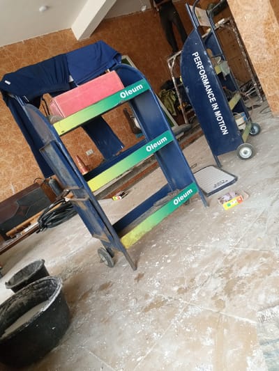 The image depicts a workshop or storage area in Hotoro North, Nigeria, characterized by a dusty, tiled floor and a light-colored ceiling. In the foreground, two large black buckets, one filled with a white substance, sit on the floor, surrounded by scattered white powder. To the right, a blue ladder with green and yellow accents stands at an angle, with the word "Oleum" visible on its steps. A red rectangular object rests on the top step of the ladder. Behind the ladder, a larger blue metal structure with wheels is visible, bearing the white text "PERFORMANCE IN MOTION". This structure appears to be some form of industrial cart or shelving unit. Further in the background, a person wearing black pants and a dark top stands on a ladder, holding an object. The walls are tiled with a terracotta-like pattern. Various other objects are scattered around, including a black hose, a dark rectangular cabinet, and what appears to be a fire extinguisher. The overall impression is of a working environment, possibly undergoing some form of construction or renovation, given the dust and scattered materials.