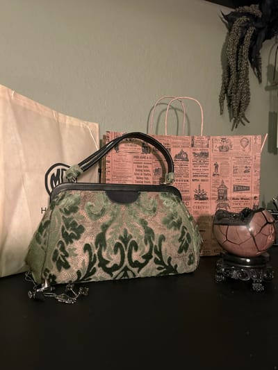 In a well-lit indoor setting, possibly a home in Cape Coral, Florida, a still life arrangement is presented. The focal point is a vintage-style handbag with a green damask pattern, featuring a black frame and handle. To the left of the handbag, a light-colored shopping bag with a circular logo partially visible is placed. Behind the handbag, two pink paper bags with a newspaper-like print adorn the background. On the right side of the composition, a cracked, reddish-pink spherical object sits on an ornate black stand. A dark, textured decorative item hangs on the wall above and to the right of the paper bags. The objects are arranged on a dark, flat surface, likely a table or shelf, against a muted green textured wall.
