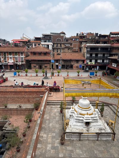 The image depicts a cityscape in Lalitpur, Nepal, with a white stupa-like structure in the foreground, partially covered by a yellow fabric-draped frame. Buildings of varying heights with red-tiled roofs and mixed architectural styles comprise the urban landscape. Potted plants line brick walls, adding greenery. People are scattered throughout the scene, some gathered near the wall in the center, others on the ground.