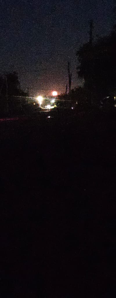 The image depicts a nighttime scene with an urban setting, possibly a street or pathway lined with trees. The foreground on the left is dark and textured, suggesting a surface like asphalt or wet pavement. Several streetlights are visible, casting a bright, diffused glow. One prominent streetlight in the mid-left emits a yellowish-white light, and further up, another is visible with a reddish-orange hue. These lights illuminate the immediate surroundings, creating reflections on the wet surface.
The background is predominantly dark, with hints of foliage from trees. The overall impression is that of a quiet, possibly slightly melancholic, urban night. There is no visible text in the image. The time of day is clearly night, and the presence of reflections suggests recent rain or a damp environment. The location is identified as Semey, Kazakhstan. The low light conditions and the grainy texture of the image contribute to a moody atmosphere. The activity, if any, is not discernible beyond the general presence of street lighting in a public area.