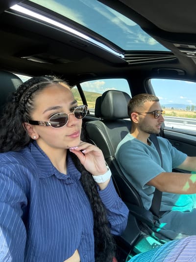 A medium shot captures two individuals, a woman and a man, seated inside a car with a sunroof open to a bright sky. The woman, positioned in the foreground on the left, wears a blue pinstriped shirt and oval sunglasses. Her right hand is near her chin, with manicured nails visible. She has braided hair. To her right, the man is driving. He wears a blue, ribbed t-shirt and sunglasses. Both individuals appear to be looking forward. The car's interior is visible, with leather seats and the central console. Outside the car, a landscape with rolling hills and trees under a blue sky with scattered clouds can be seen through the windows and sunroof. The scene suggests travel on a road in a sunny, rural environment. There is no information in the image that would place this scene specifically in the city of Kermen, Bulgaria.