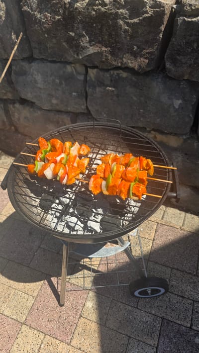 Two skewers of marinated meat and vegetables are grilling on a round charcoal barbecue. The skewers contain chunks of orange-marinated meat, white onion, and green bell pepper. The barbecue is placed on a paved patio with a stone wall in the background. The scene appears to be outdoors on a sunny day. The location is Albstadt, Germany.