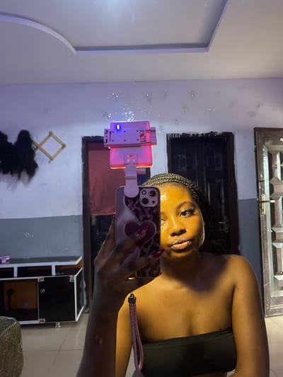 A young adult female is depicted taking a mirror selfie inside a room, presumably located in Gbongan, Nigeria. The subject, positioned centrally, has dark skin, braided dark hair, and wears a black strapless top. Her face exhibits a yellowish, possibly cosmetic, application and she has a nose piercing, with her lips slightly puckered. Her left arm is raised, holding an iPhone with a pink and white patterned case featuring a heart design. A pink braided wrist strap is attached to the phone. A clip-on ring light is affixed to the top of the phone, emitting a red glow downwards and displaying a blue indicator light.
The background, reflected in the mirror, reveals an interior room with a white ceiling featuring a decorative raised panel. The walls are light-colored, showing some texture or wear, with sparkling reflections visible, possibly from the ring light. To the left, a dark-colored wooden dresser or console table with lighter brown panels and silver legs is visible. Above it, on the wall, a light-colored wooden expanding rack holds dark, possibly fibrous, material. Two doorways are visible in the background: one partially obscured dark door frame to the left, and a more prominent dark door with a white frame and metal grating on the right side of the image. The floor appears to be tiled.
