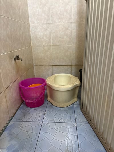 The scene depicts an interior view of a restroom facility located in Ban Hin Sam Kon, Thailand. The walls are surfaced with light beige square ceramic tiles. The floor is tiled with light blue polygonal-patterned ceramic tiles, showing some wetness. A metallic spigot is affixed to the left wall. On the floor below the spigot, a magenta plastic bucket containing an orange plastic bowl is present. To the right of the bucket, an off-white ceramic squat toilet is positioned. A corrugated metal partition, light in color, is visible on the far right, partially obscuring a dark container behind it. No individuals are present.