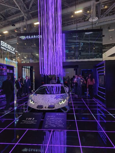 A white car is on display amidst an exhibition stand, its headlights illuminated. The backdrop features a neon grid pattern. People are gathered around the vehicle. The stand has a digital display with information on the car. Above the display, there are visible brands, "GEELY AUTO GROUP" and "CLARIOS." The exhibition is in a large venue with a high ceiling supported by metal beams.