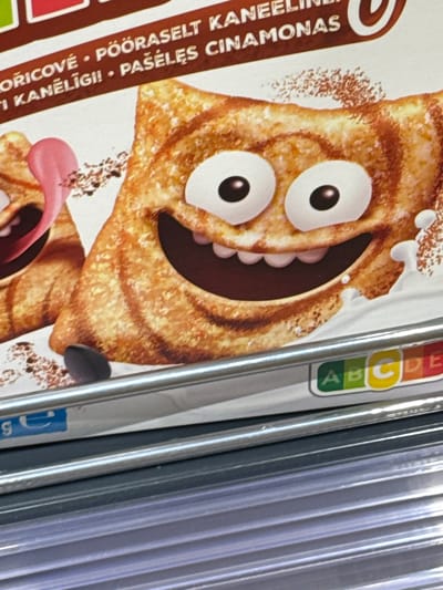 This image shows a close-up of a cereal box, featuring a cartoon character with a wide, open mouth and large eyes. The character is brown and appears to be a piece of cereal. The background is a light, textured surface. To the right of the cereal box, several thin, vertical metallic or plastic rods are visible, suggesting the box is part of a display or stored in a rack.

Visible text on the cereal box includes "ORICOVÉ · POORASELT KA", "I KANELIIGI!", and "PAŠELĖS CINAMONAS". There is also a colored bar with letters "A", "B", "C", "D", and "E" in separate colored squares (green, yellow, red, and two shades of blue). The overall impression is of a product shot or a detail of a product packaging. There are no indications of time of day, weather, or specific emotions beyond the cartoonish expression of the character. The location context is Keila, Estonia.