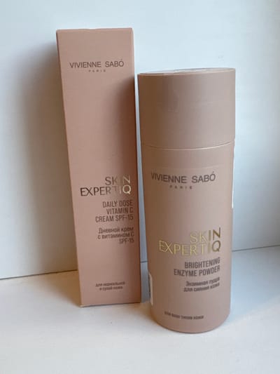 The image displays two VIVIENNE SABÓ branded cosmetic products, a box for "SKIN EXPERTIQ DAILY DOSE VITAMIN C CREAM SPF-15" and a cylindrical container labeled "SKIN EXPERTIQ BRIGHTENING ENZYME POWDER." The box is positioned to the left and slightly behind the powder container. Both items are a light peachy-pink color with gold lettering. The background appears to be a light-colored, textured surface, possibly a table or shelf, with a white wall behind. There are no people, animals, or any other distinguishing environmental features visible in the image. The lighting suggests an indoor setting with soft, diffused light. The location is not discernible from the image itself; information about Яромінскі сельскі Савет, Belarus, is external to the visual content.