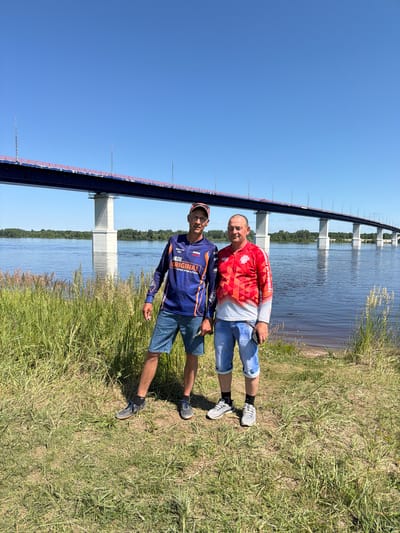 Two men are standing on a grassy bank beside a wide river on a clear, sunny day. A long, multi-span bridge with white concrete pillars crosses the river in the background. The man on the left, wearing a blue long-sleeved shirt with orange trim and denim shorts, is looking at the camera. The man on the right, wearing a red and white geometric patterned long-sleeved shirt and denim shorts, is also looking at the camera. Both men are wearing sneakers. Tall green grass and some reeds grow on the bank in the foreground. The river is calm and reflects the blue sky. The location is identified as Tarasovo, Russia.