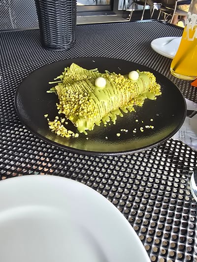 This media file depicts a close-up, overhead view of a dessert on a black plate, set against a black, perforated surface. The dessert appears to be a crepe or pastry, generously drizzled with a light green sauce and sprinkled with chopped pistachios. Two small, white spherical toppings, possibly candies or chocolates, are visible on the dessert. To the left of the plate, a portion of a black, woven object, possibly a basket or container, is visible. In the upper left corner, a yellow beverage in a clear plastic cup with a straw is partially visible. To the right, a portion of a large, white plate is present. The scene is well-lit, suggesting it is daytime. The background surface has a textured, metallic appearance. A crumpled piece of paper with some faint, unreadable text is visible behind the dessert plate. The location is identified as Divic, Bosnia and Herzegovina, though no specific landmarks or environmental cues confirm this context within the image itself. The focus is clearly on the presented dessert.