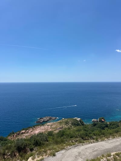 The image shows a panoramic view of the Adriatic Sea from a high vantage point overlooking the coastline near Donji Katun, Montenegro. The sky is clear and blue with a thin contrail from an airplane visible in the upper left quadrant. The sea is a deep blue, with a gentle swell. In the foreground, a dirt road curves into the frame. The coastline is rugged and covered with green vegetation, including trees and shrubs. There are several rocky outcrops in the sea, and a small red-roofed building is visible on one of them. A white boat is moving across the water in the distance. The overall impression is one of a peaceful and scenic coastal landscape on a sunny day.