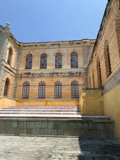The image shows a stone building with a set of stairs in front of it. The building is located in San Agustín Etla, Mexico, at the coordinates 17.197 latitude and -96.76 longitude. The building is made of yellow stone with a few rows of decorative stone on the facade. It has arched windows and a flat roof. There are five arched windows on each of the two visible stories. The building appears to be abandoned, and no people are present. There is a set of four stairs leading up to the building. The stairs are made of stone and have a slight curve. The ground in front of the building is made of stone pavers. The sky is blue and clear. The image appears to have been taken during the daytime. The building has a long, rectangular shape. The side of the building in the foreground is a solid wall with no windows or doors. The windows are not visible through the glass. There are no signs of recent activity. The image appears to be unedited and the composition is simple and straightforward. The overall mood of the image is one of calm and serenity. The image is well-lit, and there is no evidence of any motion blur. There are no visible objects in the foreground besides the stairs and the stone pavers.