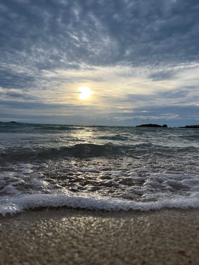 This is a vertical, close-up, eye-level shot of a beach scene at sunset, likely in Ko Samui, Thailand. The image is split into two distinct halves. On the left, the foreground shows the wet sand of the beach, glistening with moisture and reflecting the ambient light, giving it a speckled appearance. The edge of the ocean meets the sand with a foamy wave, its texture a mix of white froth and dark, turbulent water. The water itself is choppy, with sunlight reflecting off its surface in shimmering streaks.

The right half of the image is dominated by the sky. The sun, partially obscured by clouds, is low on the horizon, casting a warm, golden glow that illuminates the scene. The clouds are a mixture of light and dark gray, with streaks of white, suggesting a dynamic and possibly breezy atmosphere. The sun's rays create a bright halo effect around it. In the far distance, a dark silhouette of land, possibly an island or a prominent rock formation, is visible against the horizon.

The overall mood is serene and contemplative, capturing the peaceful transition from day to night. The lighting suggests it is either sunrise or sunset. The combination of the glistening sand, the rhythmic waves, and the dramatic sky evokes a sense of natural beauty and tranquility. No visible text is present in the image.
