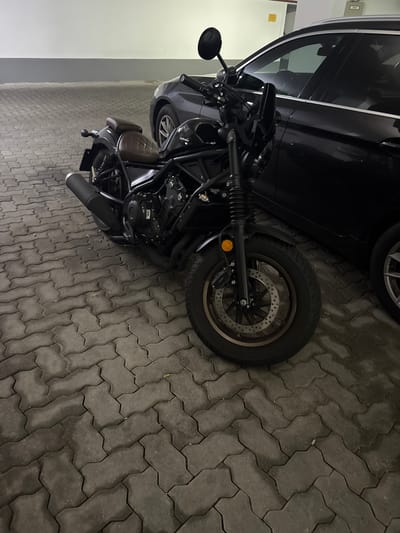 The media file displays a black motorcycle parked next to a black car. The motorcycle is angled in the frame, with its front wheel and handlebars visible on the left and its rear wheel and exhaust pipe on the right. The motorcycle has a brown seat and black detailing. The car is partially visible behind the motorcycle, with its rear wheel and side panel showing. Both vehicles are parked on a grey interlocking paver surface. The scene appears to be an outdoor parking area. The lighting suggests it could be daytime, but the overall tone is somewhat dim, possibly indicating overcast weather or an evening scene. The background to the left of the vehicles shows a light-colored wall, suggesting a building is nearby. No people are visible in the image. The visible text on the motorcycle is "NISSIN" on the brake caliper. The location context provided is Höhenkirchen-Siegertsbrunn, Germany.