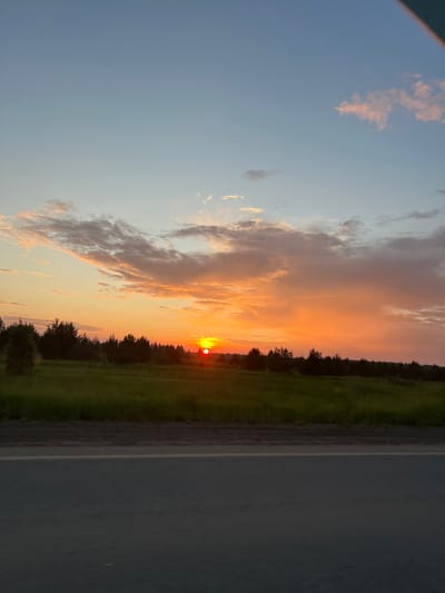 This image depicts a sunset scene in the vicinity of Поварёнки, Russia. The sky is filled with a gradient of colors, transitioning from a pale blue at the top to vibrant oranges and yellows near the horizon, illuminated by the setting sun. Wispy clouds, tinged with pink and orange, are scattered across the sky.
Below the sky, a line of dark green trees forms a dense treeline across the middle ground, suggesting a forest or woodland. In front of the treeline, a bright green field stretches out, appearing lush and slightly illuminated by the sunset.
In the foreground, a paved road runs horizontally across the bottom of the image. The road is dark gray, with a lighter gray stripe indicating a lane marker. The perspective suggests the photo was taken from a moving vehicle on this road, looking towards the sunset. No people or specific structures are visible in the scene.