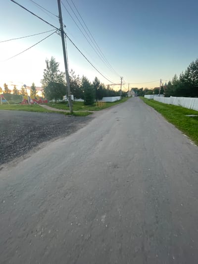 The image depicts a street scene in Bol'shoe Zaborod'e, Russia, under a clear blue sky with a hint of sunset. The foreground is dominated by an asphalt road, which appears somewhat weathered. To the left of the road, a section of dark gravel and a patch of green grass are visible, bordering a children's playground equipped with colorful structures like swings and slides. Further along the left side, trees are sparsely scattered.
The street stretches into the distance, flanked by white picket fences on both sides. Beyond the fences, houses and more trees form the backdrop. Several utility poles with numerous wires are present along the street, with one prominent pole on the left side of the road. The overall atmosphere is quiet and rural, suggesting a residential area. No people are visible in the scene.