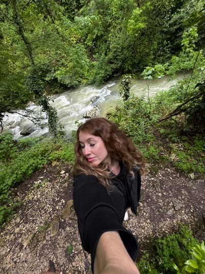 A person is standing on a dirt path beside a fast-flowing river in a forest. The river is on the left side of the image and the forest extends upwards, filling the left of the frame. The path is covered in loose rocks and vegetation, and takes up the right portion of the image. The person is wearing a black jacket and appears to be taking a selfie. The location may be in Tsabal, Abkhazia, based on information provided.