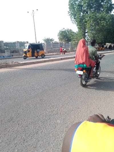 A street scene features a yellow three-wheeled vehicle near a fence, while a man in red pushes a cart in the background. In the foreground, a motorcycle carries two individuals: the driver wearing a striped shirt and a passenger draped in an orange covering. A portion of a person's arm clad in a yellow and blue shirt is visible at the bottom. The road stretches out, with a few other vehicles faintly seen in the distance. A large tree stands on the right side of the image.