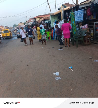 A bustling street scene is captured with several people traversing the road. A taxi, painted in yellow, is visible on the left. People stand near shops on the right, some looking at merchandise. The street itself shows signs of litter. The perspective offers a broad view of the surroundings, capturing both the human activity and the urban environment.