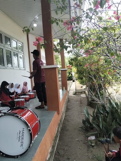 In front of a building with an orange and white facade, several young girls wearing hijabs are seated on the ground, holding drumsticks. They are arranged in a line, facing a man who is standing and appears to be instructing them. The man, wearing a maroon t-shirt with text on the back and a black baseball cap, is also holding drumsticks. Red drums are placed in front of each girl. To the right of the scene, a pathway runs alongside a lush green area with trees and pink bougainvillea flowers. The pathway is made of dirt and concrete. In the foreground, a large red drum with white accents is visible. The overall setting suggests an outdoor music lesson or practice session in Keudejungkagajah, Indonesia.
