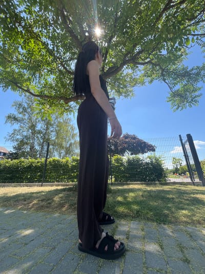 A person is standing outdoors on a sunny day, seen from a low-angle perspective looking upwards. The scene is dominated by a large tree with lush green leaves in the foreground, through which the bright sun is shining, creating a lens flare effect. The person is wearing a dark brown or black jumpsuit and black sandals. Their hair is long and dark, obscuring part of their face as they appear to be looking up or shielding their eyes. The background features a clear blue sky, a metal fence, and green grass. A paved pathway made of grey interlocking bricks is visible on the right side of the image. The lighting suggests it is daytime, likely late morning or early afternoon, given the brightness of the sun. The overall mood is relaxed and natural, set in an outdoor environment that could be a garden or park. There is no visible text in the image.