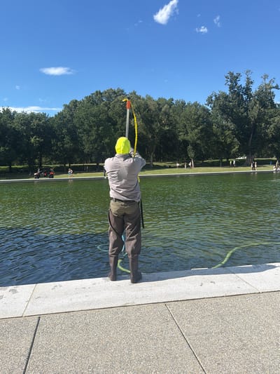 A person wearing a bright yellow hood and waders is standing on the edge of a large body of green water, holding a pole with a yellow strap attached. The person is facing away from the camera, with their back to the viewer, and appears to be working on something in the water. The person is standing on a white concrete edge, with a textured gray path in the foreground. In the background, there is a line of lush green trees, a grassy area with several people walking, and a small orange vehicle. The sky above is a clear blue with a few wispy white clouds. This scene is likely at the Tidal Basin in Washington, D.C., given the presence of the large body of water, trees, and the general park-like setting.