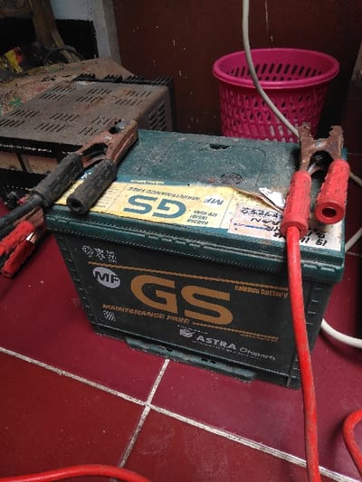 The image depicts an indoor scene in Mornington, Australia, focusing on a dark green "GS Maintenance Free calcium battery" with "ASTRA Otoparts" branding. The battery is situated on a reddish-brown tiled floor with white grout lines, which exhibit some staining and wear. A black, visibly corroded jumper cable clamp is connected to the top-left portion of the battery, with a black-sleeved cable extending leftward out of frame. A red, similarly corroded jumper cable clamp with red plastic handles is connected to the top-right, with a red cable extending downwards and out of frame. In the background, to the left, a dusty, vent-covered metallic device, possibly an old charger, rests on a surface. To the right, behind the battery, a bright pink, perforated plastic basket is visible, with a thin white cable extending towards it. The background wall appears dark. No individuals are present.