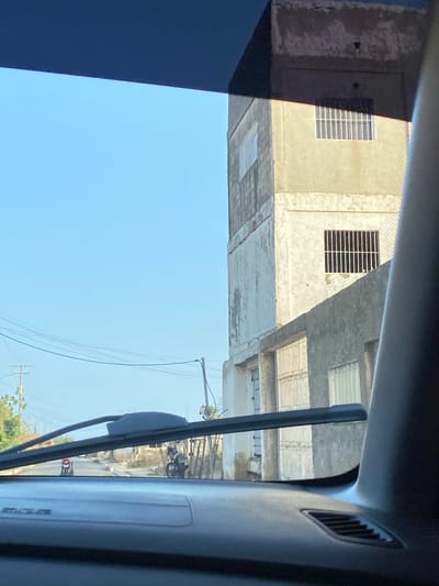 The image, taken from inside a vehicle looking out, is oriented sideways. When viewed correctly (sky up, ground down), it shows a clear, bright blue sky dominating the upper half. Below the sky, several utility poles with numerous electrical wires crisscross the frame.

The central subject consists of multi-story buildings with light-colored, possibly concrete or stucco, exteriors. Many windows feature horizontal security bars. The buildings appear somewhat worn or unfinished, with varying textures and shades of grey and beige. Sharp shadows are cast by the buildings and structural elements, indicating strong, direct sunlight, suggesting a clear day, likely mid-morning or afternoon.

In the lower portion of the image, the interior of the vehicle is visible, including a dark-colored roof liner and a side pillar on the right, which features an air vent. A windshield wiper blade is prominent on the right side, partially obscuring the view of the street level where a road and a faint outline of a small vehicle (possibly a motorcycle) can be seen.

No specific human activity is clearly discernible. No visible text is present. The architectural style, utility infrastructure, and bright conditions are consistent with an urban or semi-urban setting in La Cruz del Pastel, Venezuela.