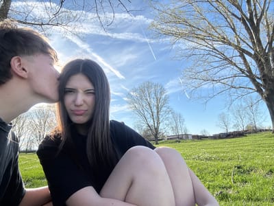 Two individuals are depicted in a photograph, oriented with the sky above and the ground below. The person on the left is frowning and has long brown hair and is wearing black clothing. The person on the right appears to be leaning in towards the left person, close to their ear.

The background shows a blue sky with white contrails. Bare tree branches are visible. The ground is visible in the top left of the image and appears to be grassy. The location is in Beli Brod, Bulgaria.