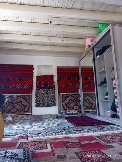 The image shows an indoor scene, likely a bedroom, with traditional carpets and textiles. In the foreground, several layered carpets with intricate geometric patterns are laid out on the floor. The colors are predominantly red, pink, and blue, with white and black accents. Behind the carpets, to the left, are two tall, narrow cabinets with ornate carved wooden doors, each displaying folded red and patterned textiles stacked within recessed shelves. To the right of these is a large, modern wardrobe with mirrored doors, revealing shelves containing folded clothes, bags, and other items. Plush toys, a green frog and a pink pig, are visible on top of the wardrobe. The ceiling is made of exposed white-painted wooden beams. A simple light fixture hangs from the ceiling. The overall impression is one of a densely decorated and lived-in space, possibly a guest room or a living area, with a blend of traditional and modern furnishings. The timestamp at the bottom right corner indicates "30.06.2026 11:31". The scene is consistent with domestic interiors found in Batken City, Kyrgyzstan, known for its rich cultural heritage and traditional crafts, including carpet weaving.