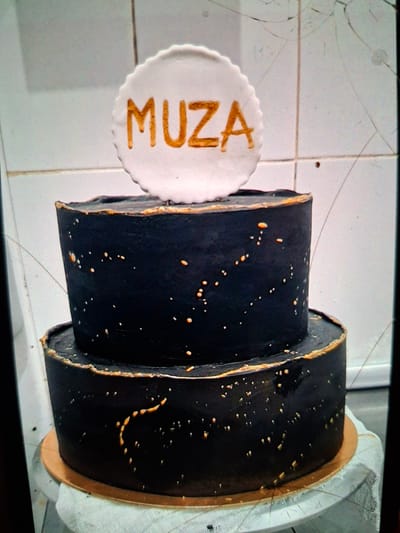 A two-tiered black cake is presented on a light-colored turntable covered with a white cloth. The cake is decorated with small, irregular gold splatters, resembling stars in a night sky, and a gold trim around the edges of each tier. Atop the cake, a white circular plaque with a scalloped edge displays the word "MUZA" in gold lettering. The background appears to be a tiled surface, possibly in a kitchen or bakery setting. The image is taken from a slightly elevated angle, and there are visible cracks on the surface where the photograph was taken, possibly a screen or glass. The location is noted as Medvezhyi Ozyora, Russia.
