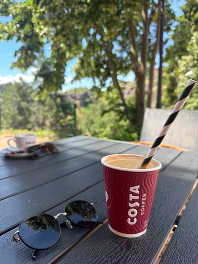 The image depicts a close-up view of a table outdoors, likely at a cafe, with a Costa Coffee cup and a pair of sunglasses in the foreground. The table appears to be made of dark, slatted wood. To the left of the Costa cup, there is a white ceramic mug and saucer, also on the table. A black and white striped straw is inserted into the Costa Coffee cup. The background is blurred, showing green foliage and trees against a bright sky, suggesting a sunny day and a natural or park-like setting. The overall atmosphere is relaxed and casual. The provided context indicates that the location is the city of Belogradchik, Bulgaria.