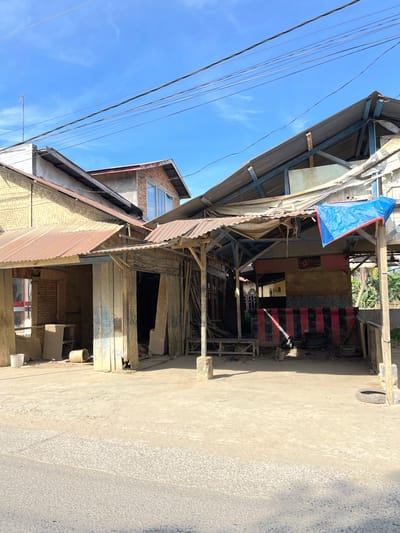 In Kota Kuala Simpang, Indonesia, this photograph captures a street scene featuring a collection of structures with rustic, possibly unfinished or dilapidated, roofing and walls. The buildings appear to be a mix of residential and commercial, with some displaying corrugated metal roofs and others featuring woven bamboo walls. In the foreground, a dirt or gravel road is visible. The sky is a clear blue with wispy clouds, and overhead, power lines crisscross the frame. The structures themselves are characterized by weathered wood, exposed beams, and various materials used for roofing and siding. One structure has a prominent blue tarp draped over a portion of its roof, and another has a striking red and black striped pattern on its facade. Several objects are scattered in front of the buildings, including buckets, tires, and wooden planks, suggesting ongoing construction, repair, or storage. The overall impression is one of a functional, lived-in environment, possibly in a developing or rural part of the city.