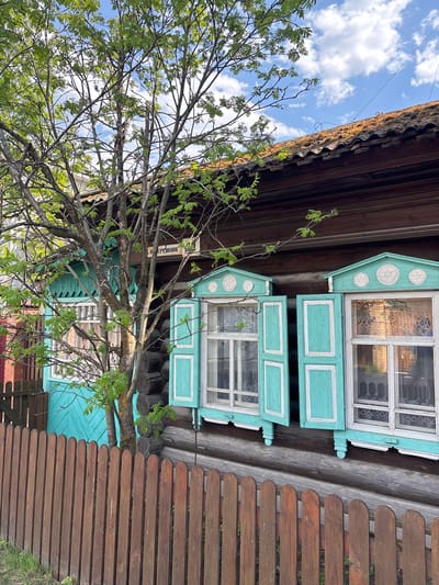 The image depicts a traditional wooden house in the city of Овсянка, Russia, partially obscured by a tree. The house features two windows with ornate turquoise and white painted frames. A brown wooden fence stands adjacent to the house. The sky is visible through the tree branches, with scattered clouds.