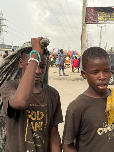 The image displays two young males in the foreground of a street market environment in Jakande, Nigeria, under an overcast sky. The male on the left, with short dark hair, wears a dark brown t-shirt featuring faded gold text, "GOD MAN! believe...". His left arm is raised, with his hand gripping a bundled, dark, textured fabric that is draped over his head and shoulder. His left wrist bears a white beaded bracelet and a turquoise spiral bracelet. His head is angled downwards, obscuring his direct gaze. The male on the right, also with short dark hair, wears a dark brown t-shirt, and a crumpled, yellowish, fibrous cloth is draped over his right shoulder. He faces slightly right, with his mouth slightly ajar.
In the midground, a bustling market scene is visible on an uneven ground surface. Several individuals are present, including a man in a light grey traditional outfit standing near colorful market umbrellas (red, blue) that shade various goods, possibly liquids in plastic bottles. Another person in a red shirt is seated on a stool. A dark vehicle is partially visible on the far right. A tall utility pole on the right side of the frame features a large commercial banner advertising a "PHOTO STUDIO" with contact numbers. The background is dominated by multiple large metal electricity pylons and a network of high-voltage power lines against the cloudy sky.