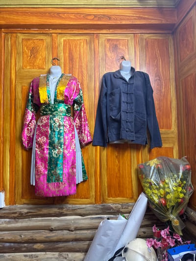 The image, likely taken in Mèo Vạc Commune, Vietnam, shows a scene with several objects placed on a wooden surface that appears to be a wall or possibly a door.
A dark blue jacket is presented on a white, partial mannequin. Below it, there's a colorful garment, possibly a traditional dress or robe, also displayed on a partial mannequin. This item features a vibrant combination of pink, green, and gold fabrics with an intricate floral pattern.
To the upper right, a plastic bag contains yellow and red flowers. Adjacent to this are other items: a white helmet, documents or rolled paper labeled "SAILS", pink decorative flowers, and a blue shopping bag.
The background consists of a wooden surface that varies from the painted wall behind the clothing to the raw, unfinished wood.