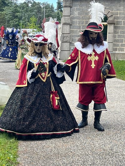 Two individuals in elaborate historical costumes stand on a gray stone surface, likely a walkway. The costumes evoke a period style, possibly 17th or 18th century, with elements like lace collars, rich fabrics, and decorative crosses.
One costume is primarily red with gold trim and features a masked character wearing a feathered hat and black boots. The other costume incorporates a dark, patterned fabric with gold and red accents and a long, elaborate skirt. The person is also wearing a mask and holds a fan.
In the background, other costumed figures can be seen, indicating an event or festival. Trees, a building, and overcast sky suggest an outdoor setting, likely in Saint-Galmier, France. The presence of other people in costumes, umbrellas, and other details add context to the scene.