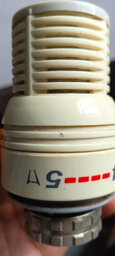 This is a close-up, high-angle, eye-level, medium shot of a cream-colored object, possibly a dial or knob, with blue and red markings. The object is dirty and has some scuff marks. The top of the object is ribbed and appears to be made of plastic. Below the ribbed section is a smooth, rounded section. Further down, there is a cylindrical section with markings. A blue character that resembles the letter "A" or a triangle is to the left of a large blue "5". To the right of the "5" is a series of red dashes. The bottom of the object has a knurled metal ring, suggesting it is a component that can be turned. The background is out of focus and appears to be a light gray. The object is held by a hand, partially visible at the bottom of the frame. There are no discernible elements that would allow for localization to Tecnimont camp, Russia.