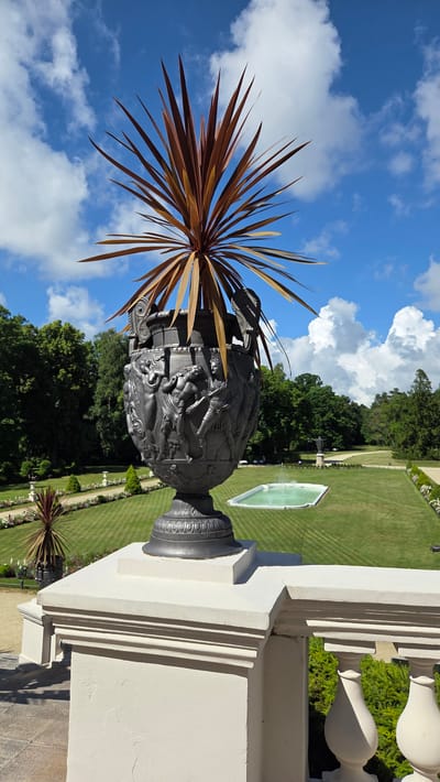 A large, dark, ornate urn stands prominently in the foreground, filled with a spiky, reddish-brown plant. The urn is intricately decorated with bas-relief carvings depicting figures engaged in what appears to be a mythological scene. It rests on a white stone balustrade, suggesting it's part of a terrace or balcony.

Beyond the urn and balustrade lies a meticulously manicured garden. Rolling green lawns are striped with subtle mowing patterns, and rows of flowerbeds line pathways. In the middle ground, a rectangular pool with turquoise water sits surrounded by grass. Further into the background, a dense line of mature trees forms the edge of the estate, under a bright blue sky dotted with fluffy white clouds. To the left, a smaller, similar urn with a spiky plant is visible in the distance.

This scene is located in Palanga, Lithuania, likely within the grounds of the Palanga Botanical Garden or a similar grand estate. There are no people visible in the image, and no actions or interactions are taking place. The overall atmosphere is one of serene, well-maintained grandeur.