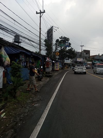 This image depicts an active street scene under an overcast, gray sky. Visual evidence from signage, specifically the text "BADRUM" and "SELALU PAS", indicates the location is an Indonesian-speaking region, contradicting the provided location of Dawathang_Dorjibi_ Kashingtsawa, Bhutan.

On the left side of the frame, a row of makeshift roadside stalls or small shops are visible. These structures feature blue tarpaulin coverings and corrugated metal roofs. A black water tank is present on the roof of the leftmost stall. Merchandise, including items in clear plastic bags, hangs from the stalls or is displayed on rudimentary shelves. Adjacent to these stalls, a male individual is standing on the unpaved ground, wearing a dark short-sleeved shirt, light-colored shorts, and dark sandals. He is positioned near a dark-colored scooter parked on the dirt. Small green plants are present along the immediate roadside. A prominent wooden utility pole heavily laden with a dense network of black electrical and communication cables dominates the upper left and central portions of the scene.

The road itself is a dark asphalt surface, curving gently to the right as it extends into the background. A solid white line delineates the edge of the road from the unpaved shoulder. Further down the road, a double yellow line marks the center. Several vehicles are in motion: a light-colored SUV with roof racks is visible heading away from the camera in the lane furthest to the right, followed by another vehicle. A light-colored van is visible in the opposing lane, approaching the camera.

In the mid-distance and background on the right side of the road, various buildings of different heights are visible. Several trees with green foliage are interspersed. A tall, empty metal framework for a billboard stands behind the main utility pole. Road signs are also present, including a blue rectangular sign with white text (possibly "BADRUM"), another blue sign with a mosque symbol, and a yellow diamond-shaped pedestrian crossing warning sign. The overall lighting is diffused, consistent with an overcast day.